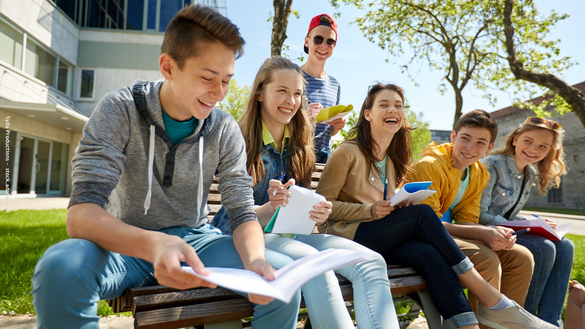 Un groupe d’adolescents souriants assis sur un banc à l’école, partageant leurs cahiers et riant ensemble, symbole d’amitiés et de socialisation à la rentrée.