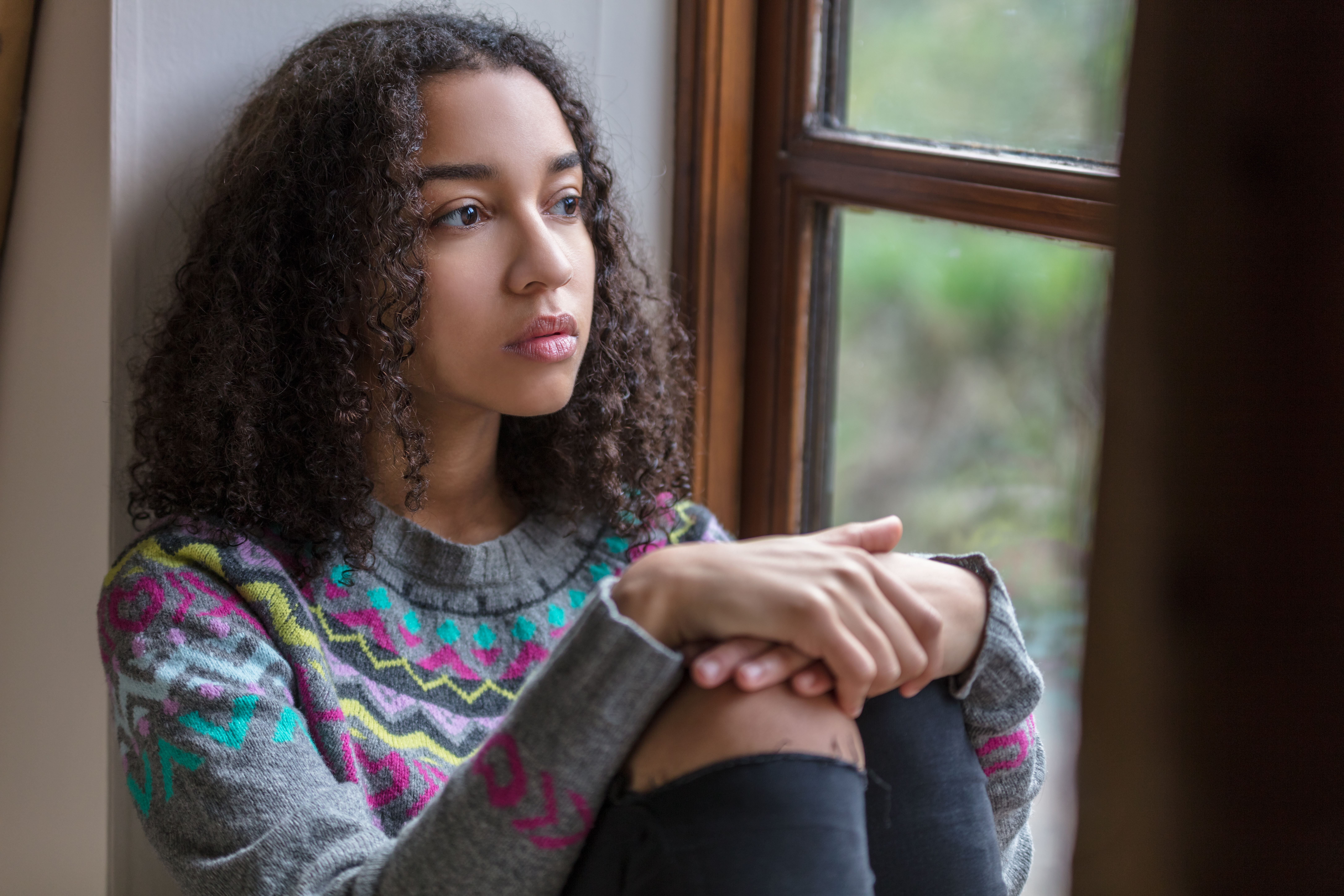 Adolescente pensive au regard triste assise près d'une fenêtre, genoux repliés contre elle