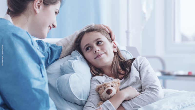 Une fillette hospitalisée sourit à une soignante bienveillante, illustrant le soutien et l’accompagnement des enfants malades et de leurs parents.