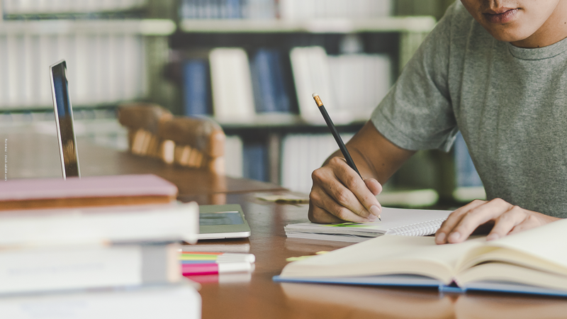 Élève concentré à son bureau, écrivant dans un cahier entouré de livres : illustration du travail scolaire pendant les vacances.