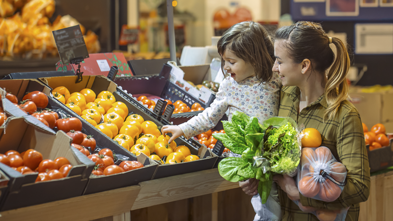 Une mère et sa fille choisissent des légumes au marché, illustrant la vie quotidienne et l’équilibre budgétaire d’une famille monoparentale.
