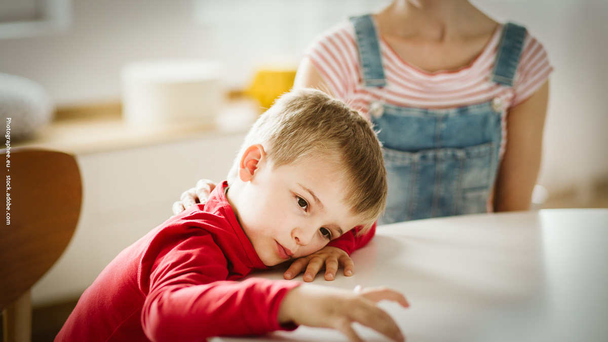 Un jeune garçon en pull rouge, l’air pensif et fatigué, appuyé sur une table, avec une adulte bienveillante à ses côtés. Image illustrant les difficultés scolaires ou le mal-être lié aux troubles DYS, TDAH ou TSA.