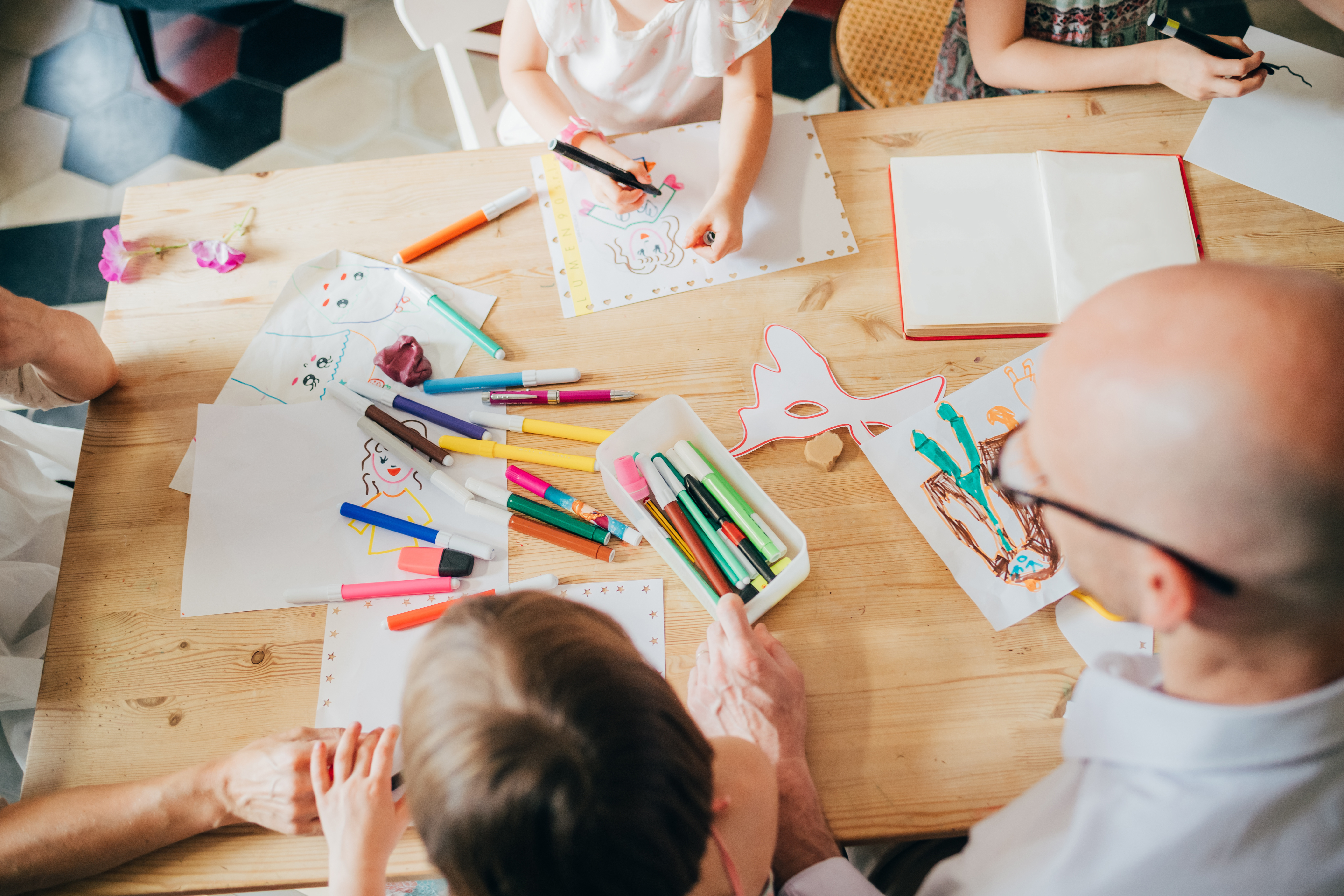 telier créatif en famille avec enfants et adultes dessinant et coloriant des masques et personnages à la table, entourés de feutres.
