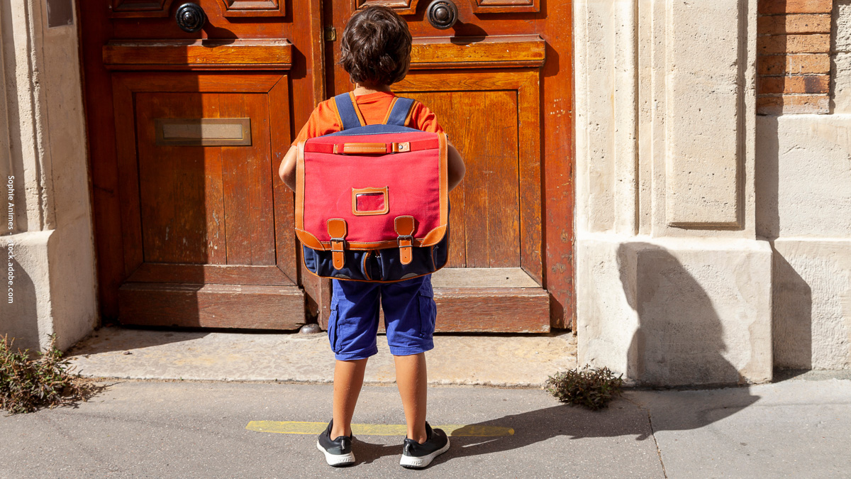 Un enfant avec un grand cartable rouge se tient devant la porte de son école, prêt pour sa première rentrée en CP. Une étape symbolique pleine d’émotions.