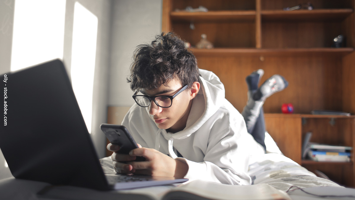 Collégien allongé sur son lit utilisant son smartphone à côté d’un ordinateur, illustrant l’usage du téléphone portable à l’entrée en 6ᵉ.