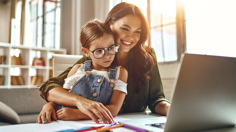 Une mère souriante travaille sur un ordinateur avec sa fille sur les genoux, illustrant la conciliation entre emploi et vie de parent solo.