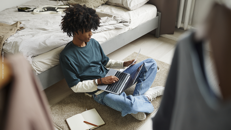 Adolescent assis au sol dans sa chambre, adossé au lit, utilisant un ordinateur portable