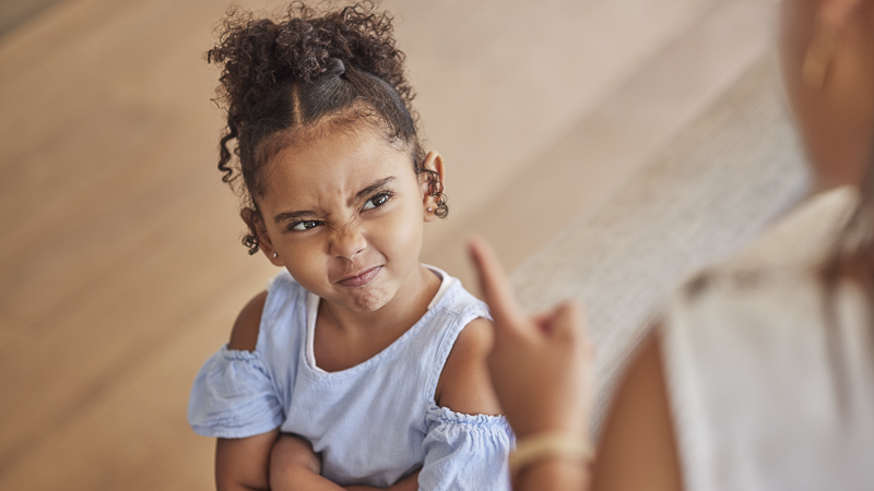 Petite fille les bras croisés, air buté, regardant un adulte pointer le doigt vers elle — enfant qui n'écoute pas