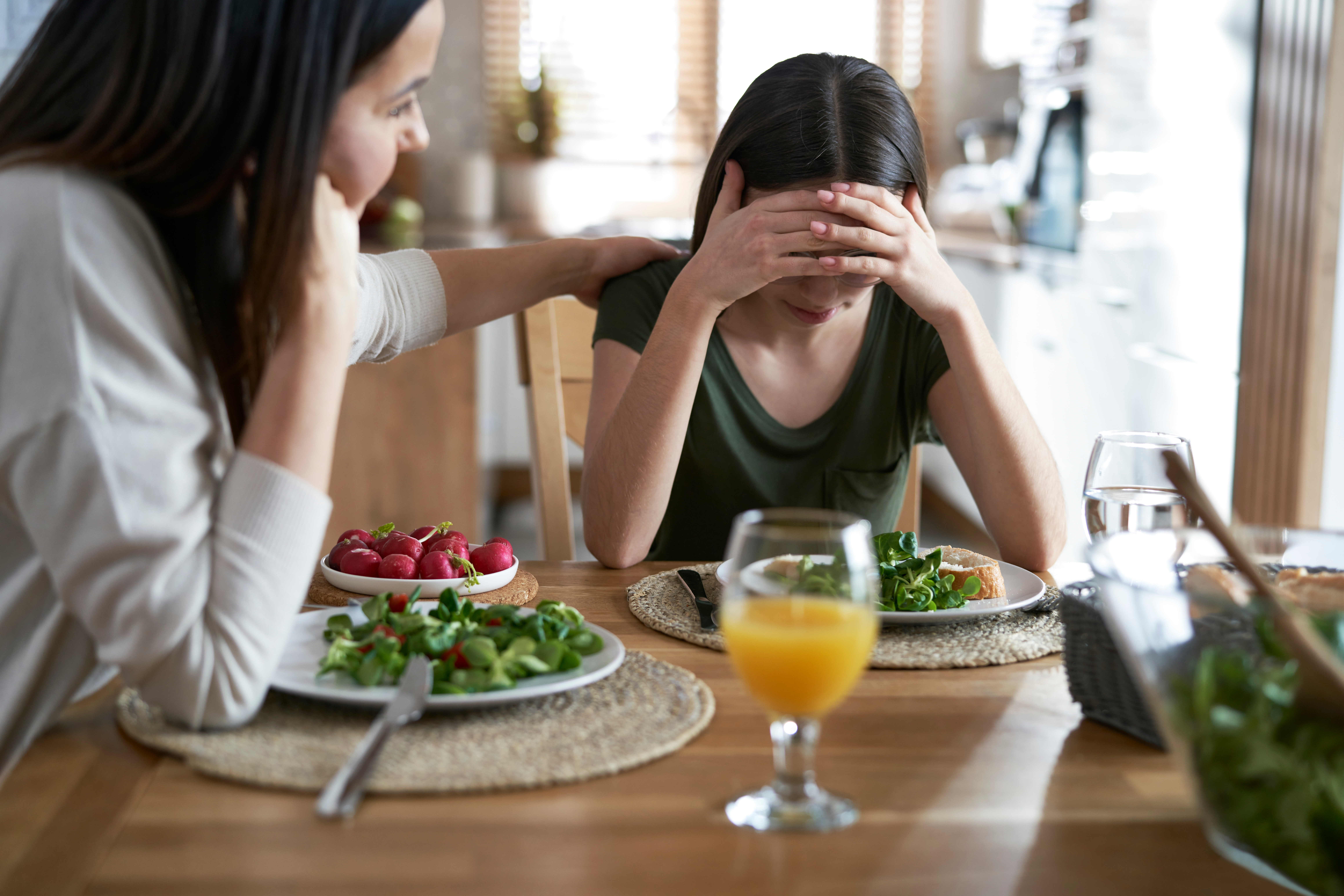 Adolescente assise à table cachant son visage pendant un repas, tandis que sa mère tente de la réconforter.