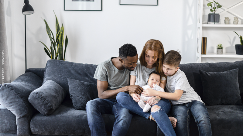Famille recomposée assise sur un canapé, parents et enfants réunis autour d’un bébé, partageant un moment de tendresse et de complicité.