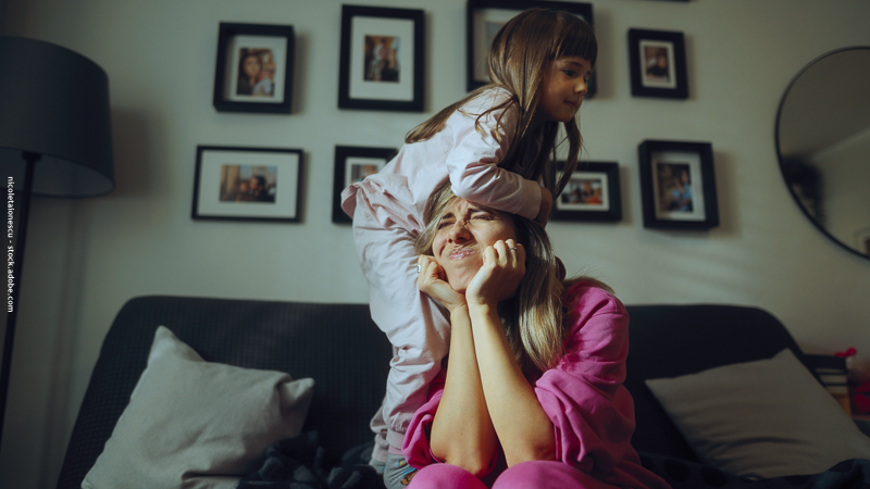 Mère assise sur un canapé, air exaspéré, pendant que sa fille se tient debout derrière elle, illustrant les tensions et le manque d’écoute à la maison.