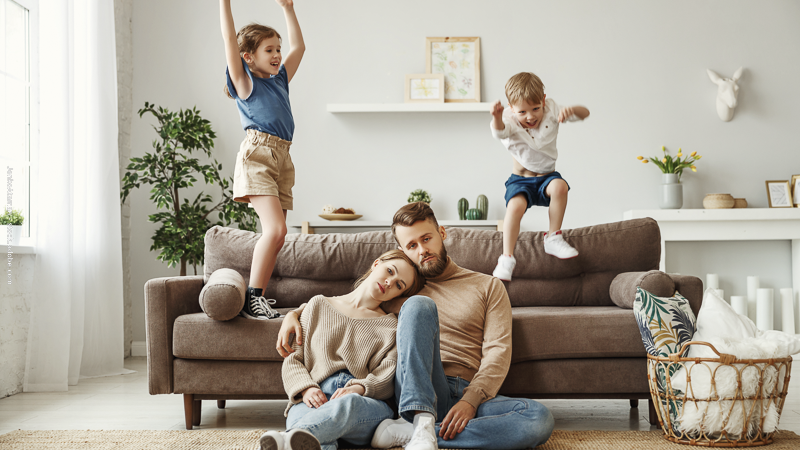 Parents épuisés assis au sol devant un canapé tandis que leurs enfants sautent dessus, illustrant la fatigue et la surcharge parentale.