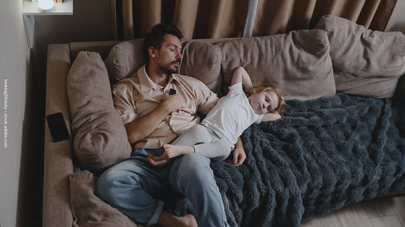Père endormi sur un canapé pendant que son enfant dort sur lui, illustrant la fatigue parentale en fin de journée.